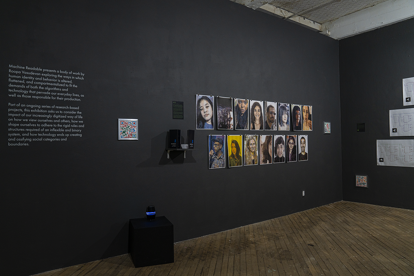 A wide view of a gallery with gray walls and wood floors. On the left side of the image there are 15 unframed prints hung up on the wall of people's portraits, and 2 small framed prints with QR codes on either side of them. On the right side is another small QR code print, hung close to the ground, and three large, unframed white systems diagrams tacked next to it.